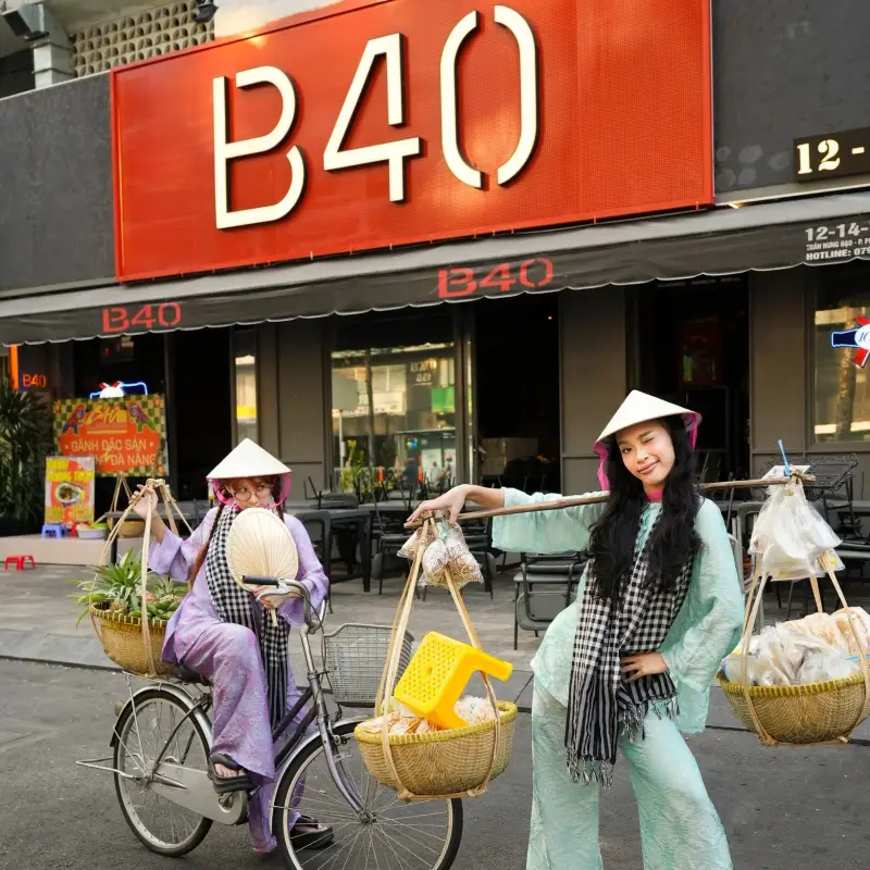 Hai cô gái mặc áo dài, đội nón lá với quang gánh và xe đạp trước cửa B40